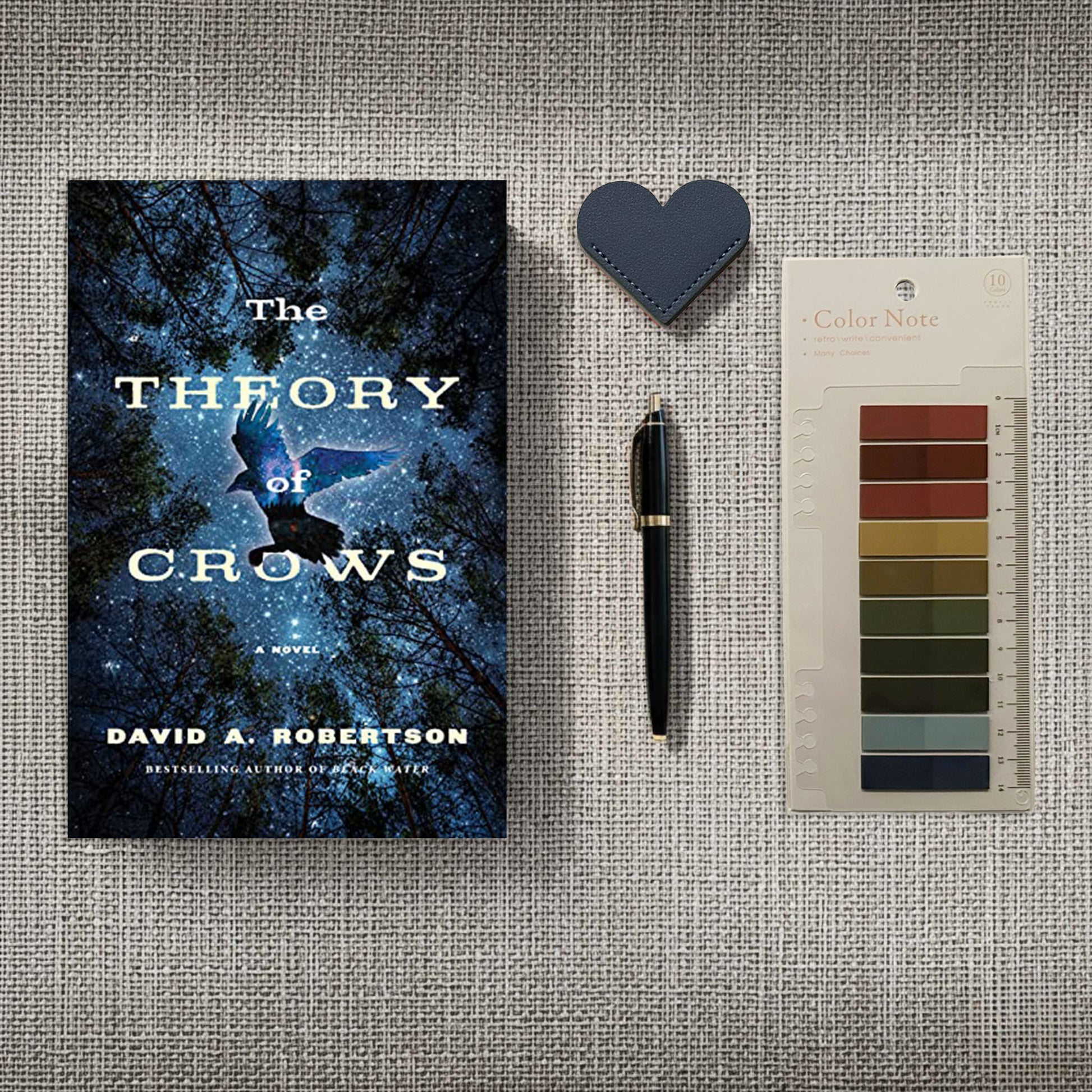 The novel The Theory of Crows anchors the scene, its cover art of a crow soaring through mist and starlight perfectly capturing the theme's mystical energy. Accompanied by a deep obsidian-blue leather heart bookmark and "Color Note" tabs in earthy, heather-inspired tones, this arrangement feels like a curated set of tools for deciphering forgotten lore in a sun-drenched, fog-shadowed study.