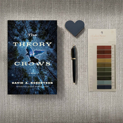 The novel The Theory of Crows anchors the scene, its cover art of a crow soaring through mist and starlight perfectly capturing the theme's mystical energy. Accompanied by a deep obsidian-blue leather heart bookmark and "Color Note" tabs in earthy, heather-inspired tones, this arrangement feels like a curated set of tools for deciphering forgotten lore in a sun-drenched, fog-shadowed study.