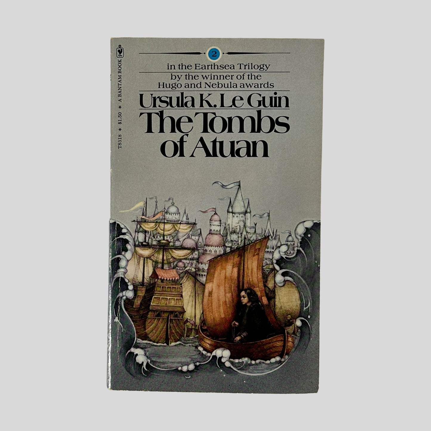 The front cover of the Bantam edition of The Tombs of Atuan by Ursula K. Le Guin, featuring a detailed illustration of a dark-haired figure in a boat near a majestic seaside castle.