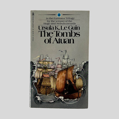 The front cover of the Bantam edition of The Tombs of Atuan by Ursula K. Le Guin, featuring a detailed illustration of a dark-haired figure in a boat near a majestic seaside castle.