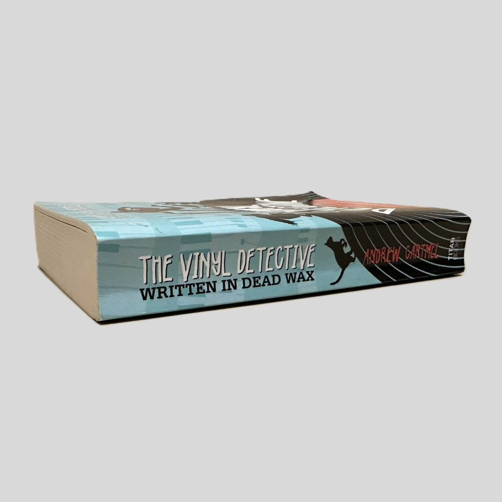 Close-up spine view of The Vinyl Detective by Andrew Cartmel centered on a neutral light gray background. Spine shows creasing. Pages lay flat.