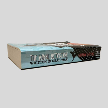 Close-up spine view of The Vinyl Detective by Andrew Cartmel centered on a neutral light gray background. Spine shows creasing. Pages lay flat.