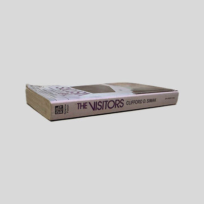 Close-up spine view of The Visitors by Clifford D. Simak centered on a neutral light gray background. Spine shows creasing. Pages lay flat.