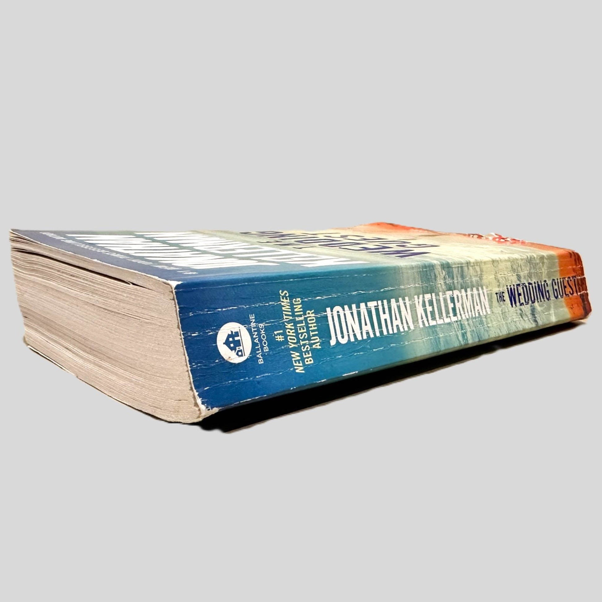 Close-up spine view of The Wedding Guest by Jonathan Kellerman centered on a neutral light gray background. Spine shows creasing and cocking. Pages lay flat with the exception of a dog-eared page.