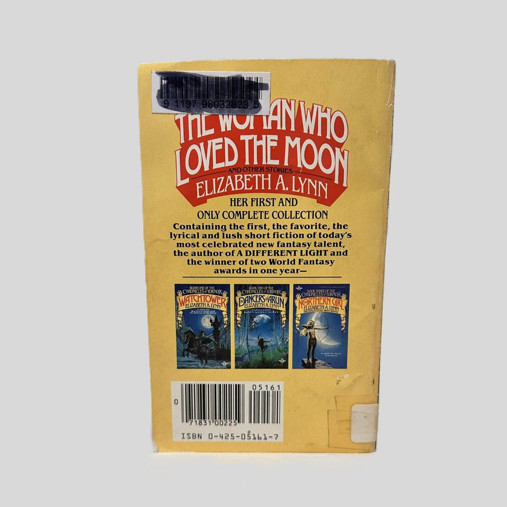Back cover view of The Woman Who Loved The Moon by Elizabeth A. Lynn centered on a neutral light gray background. Library barcode sticker in top left corner, colored over with black ink. Chipping and creasing are present around the edges.