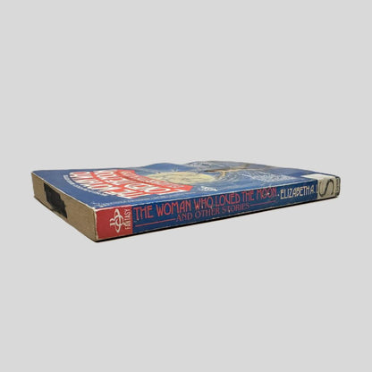 Close-up spine view of The Woman Who Loved The Moon by Elizabeth A. Lynn centered on a neutral light gray background. Spine is square and shows minimal creasing. Wave is present toward the page edge otherwise the pages lay flat.