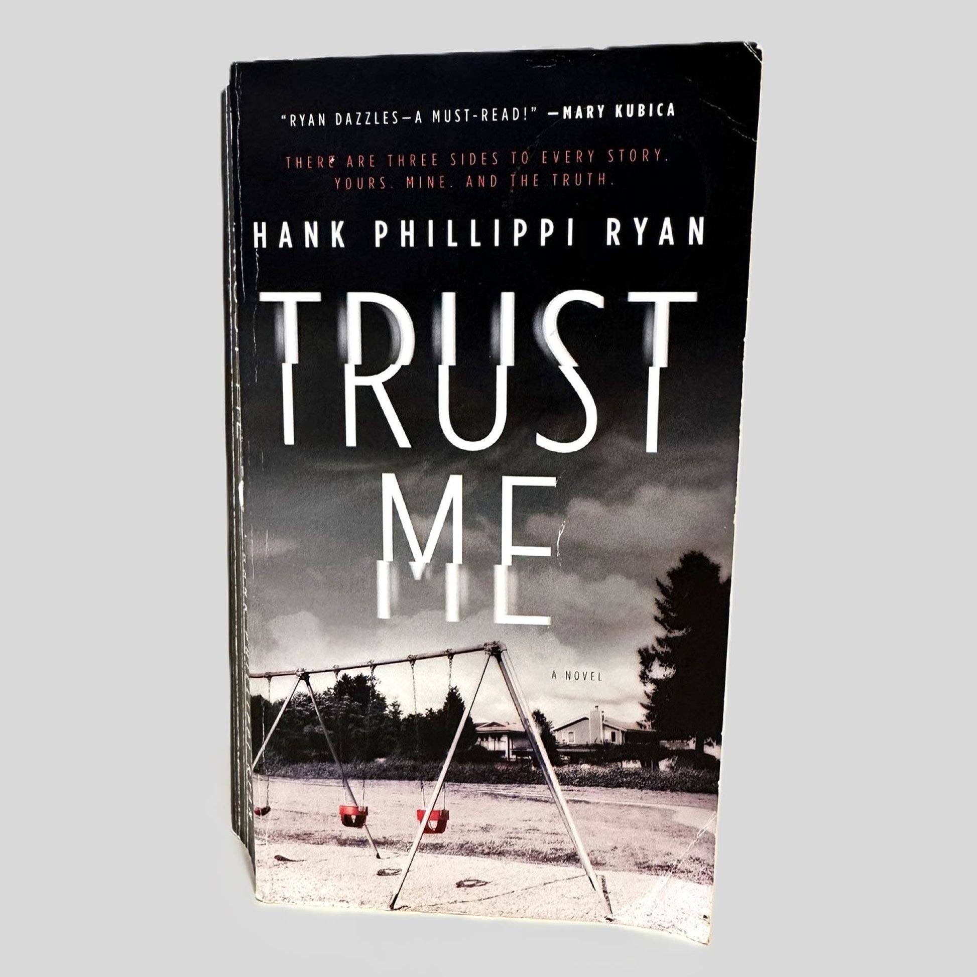 Front cover view of Trust Me by Hank Phillippi Ryan centered on a neutral light gray background. Chipping is present along the edges of the cover.