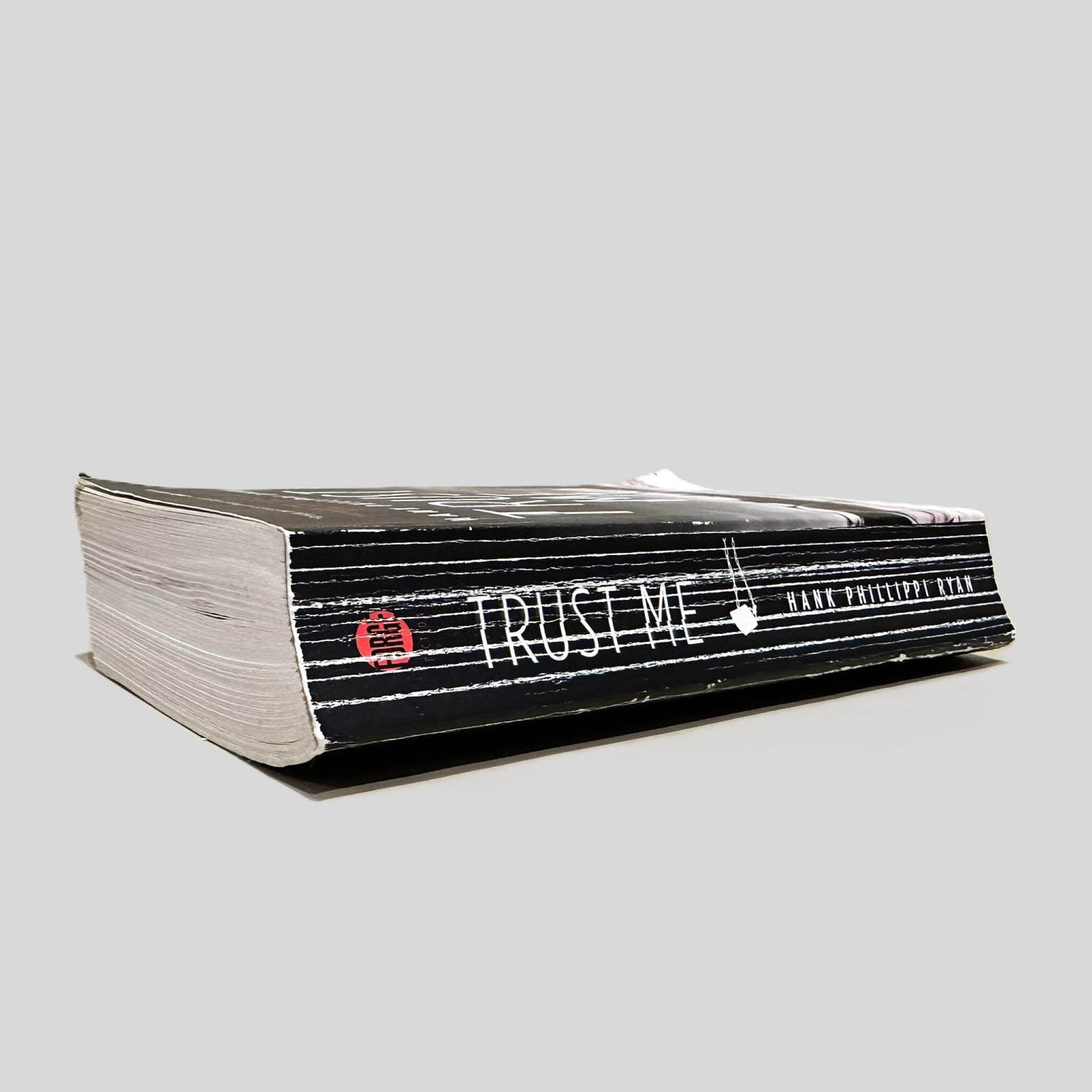 Close-up spine view of Trust Me by Hank Phillippi Ryan centered on a neutral light gray background. Spine shows creasing and cocking. Pages lay flat.