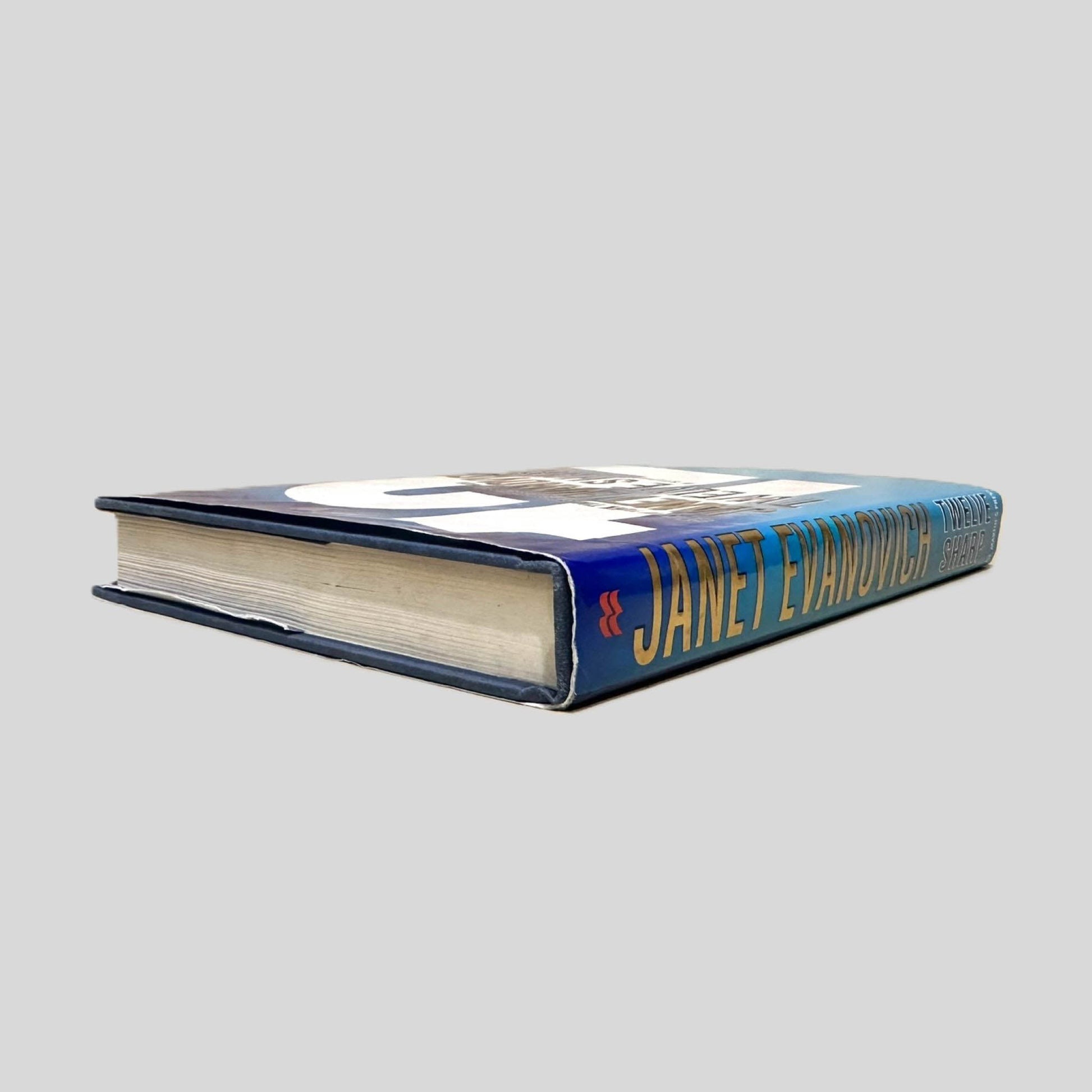 Close-up spine view of Twelve Sharp by Janet Evanovich centered on a neutral light gray background. Spine shows no creasing. Pages lay flat.