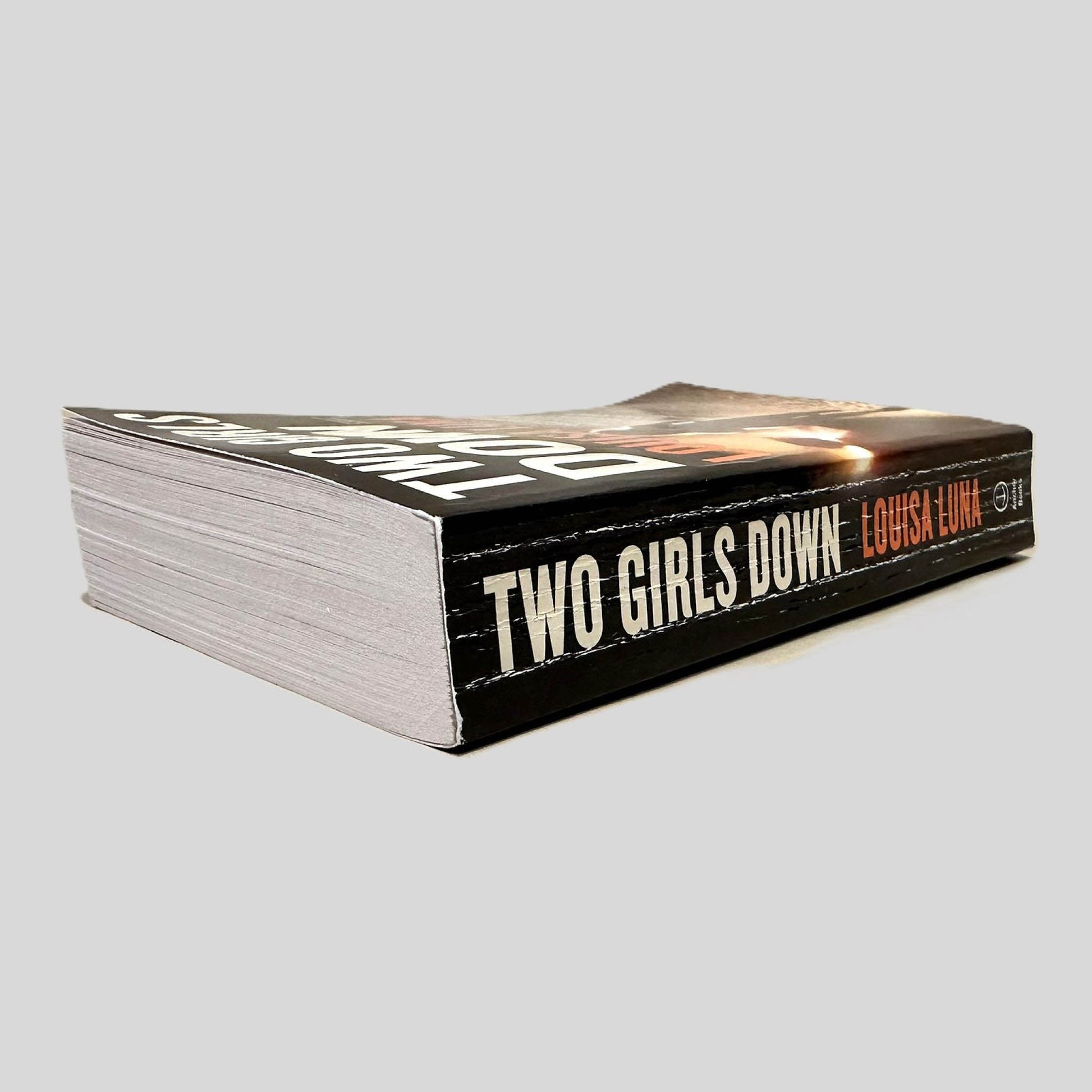 Close-up spine view of Two Girls Down by Louisa Luna centered on a neutral light gray background. Spine is square, shows creasing. Pages lay flat.