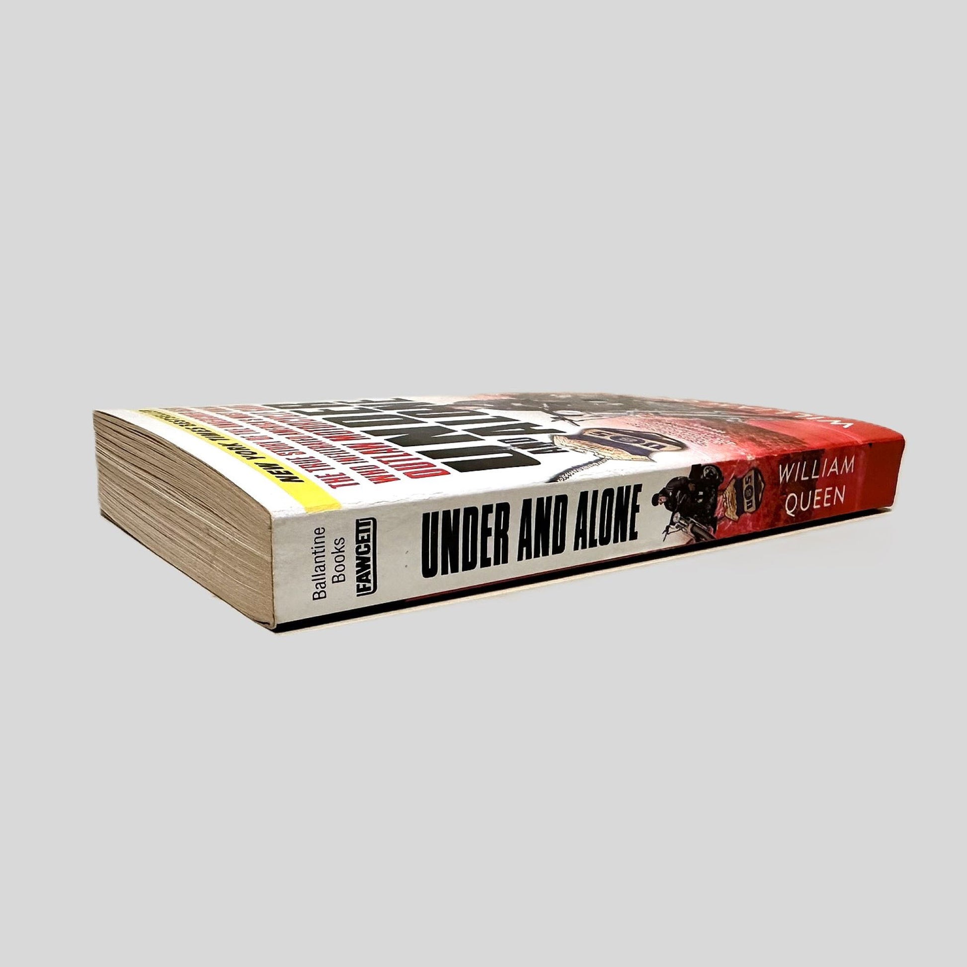 Close-up spine view of Under and Alone by William Queen centered on a neutral light gray background. Spine is square and shows minimal creasing.