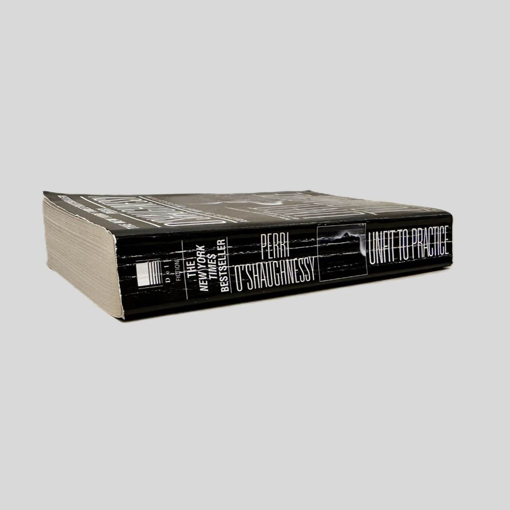 Close-up spine view of Unfit to Practice by Perri O'Shaughnessy centered on a neutral light gray background. Spine shows creasing.