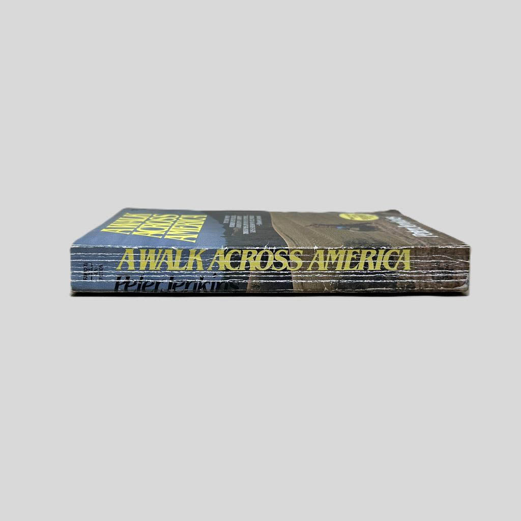 Close-up spine view of Walk Across America by Peter Jenkins centered on a neutral light gray background. Spine shows creasing and chipping in the lower corner of the spine. Pages lay flat.