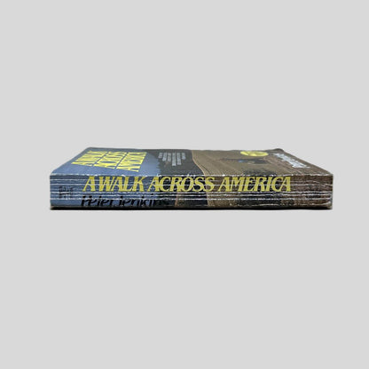 Close-up spine view of Walk Across America by Peter Jenkins centered on a neutral light gray background. Spine shows creasing and chipping in the lower corner of the spine. Pages lay flat.