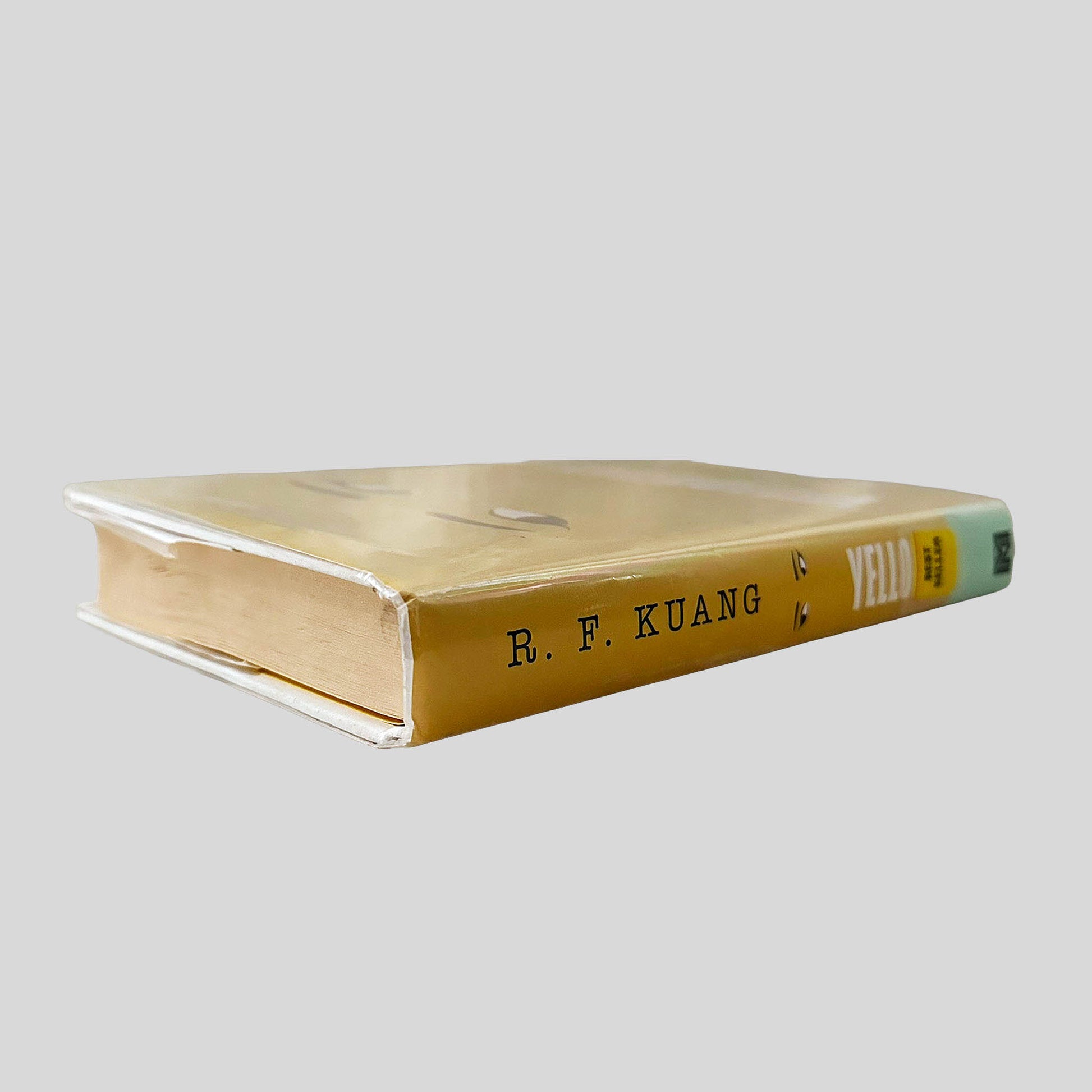 Close-up spine view of Yellowface by R. F. Kuang centered on a neutral light gray background. Dust jacket is present and pristine, wrapped in a mylar cover. Pages lay flat.