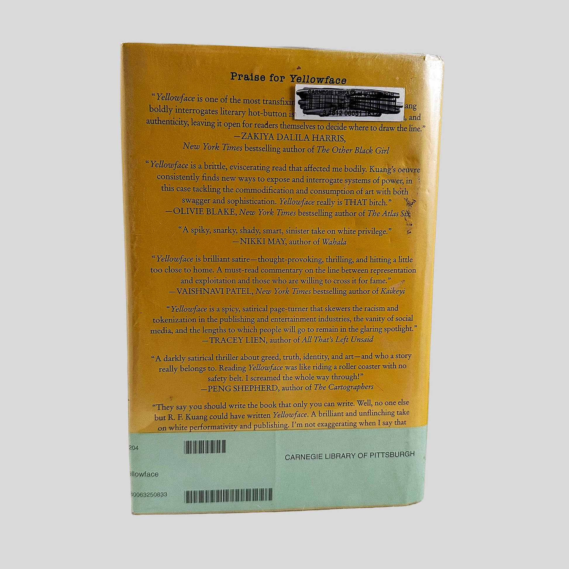 Back cover view of Yellowface by R. F. Kuang centered on a neutral light gray background. Dust jacket is present, in a mylar cover. Library barcode sticker in the top right, covered with black marker.
