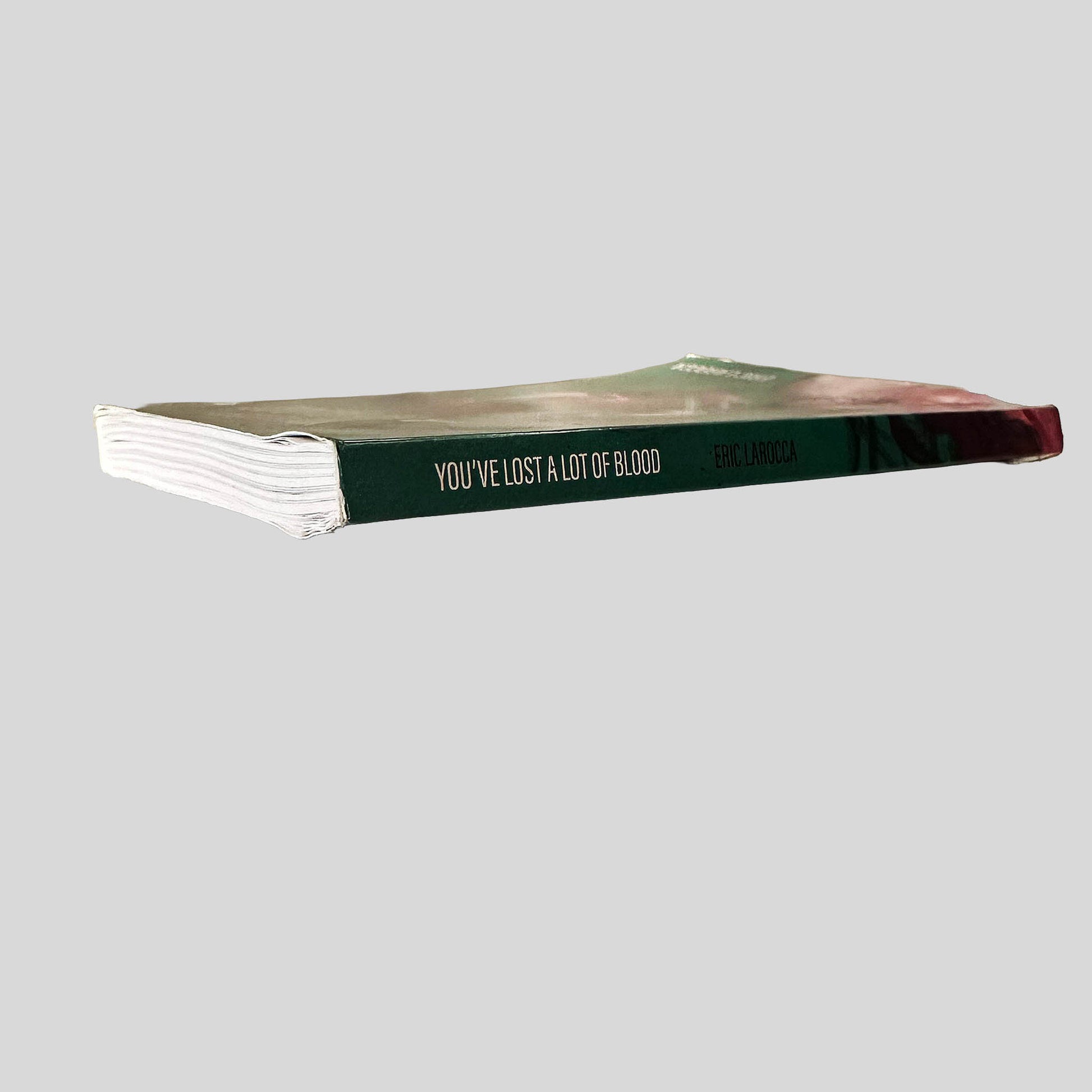 Close-up spine view of You've Lost A Lot of Blood by Eric LaRocca centered on a neutral light gray background. Spine shows minimal creasing. Pages wave at the spine before laying flat.