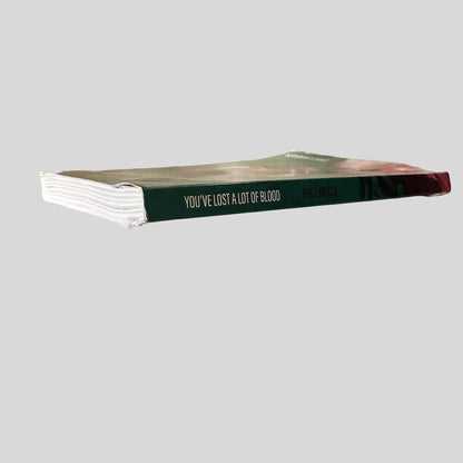 Close-up spine view of You've Lost A Lot of Blood by Eric LaRocca centered on a neutral light gray background. Spine shows minimal creasing. Pages wave at the spine before laying flat.