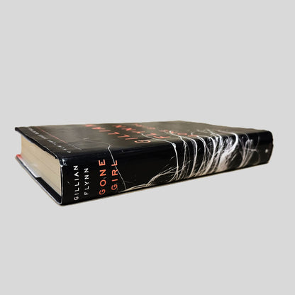 Side-angle view of 'Gone Girl' by Gillian Flynn resting on a neutral background. The screen captures the striking title and Crown Publishers logo on the spine, reflecting the high-quality design of this bestselling first edition. The layout emphasizes the chilling atmosphere of the novel, showcasing the sharp corners and authentic dust jacket of this collectible hardcover volume.