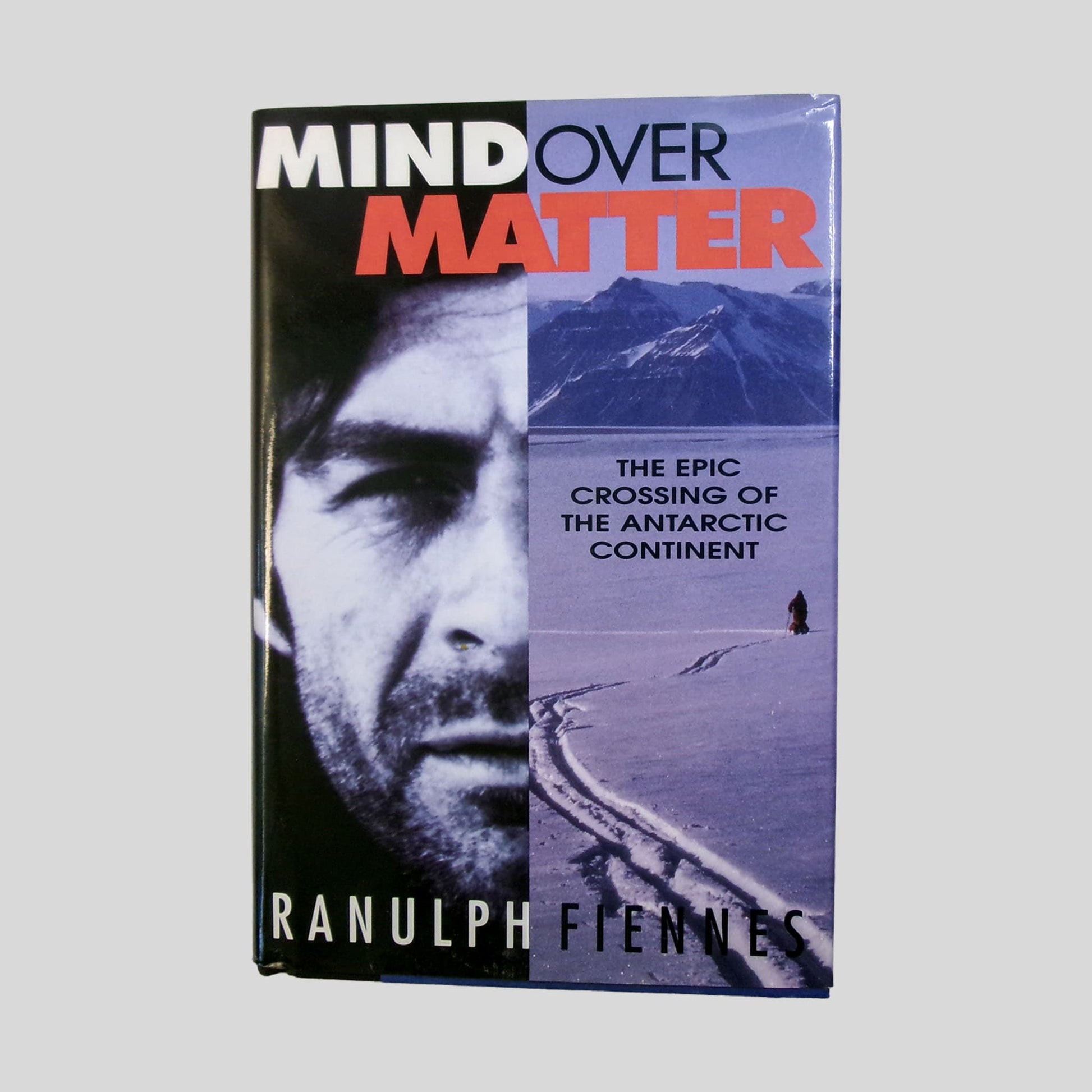 The front cover of "Mind Over Matter" features a stark black-and-white portrait of Ranulph Fiennes juxtaposed against a cold blue Antarctic landscape. The screen showcases the bold red and white typography of the title and the subtitle, "The Epic Crossing of the Antarctic Continent," alongside a lone figure on a vast field of ice. This Fehmerling Books hardcover is a premier addition for readers of survival and adventure memoirs.