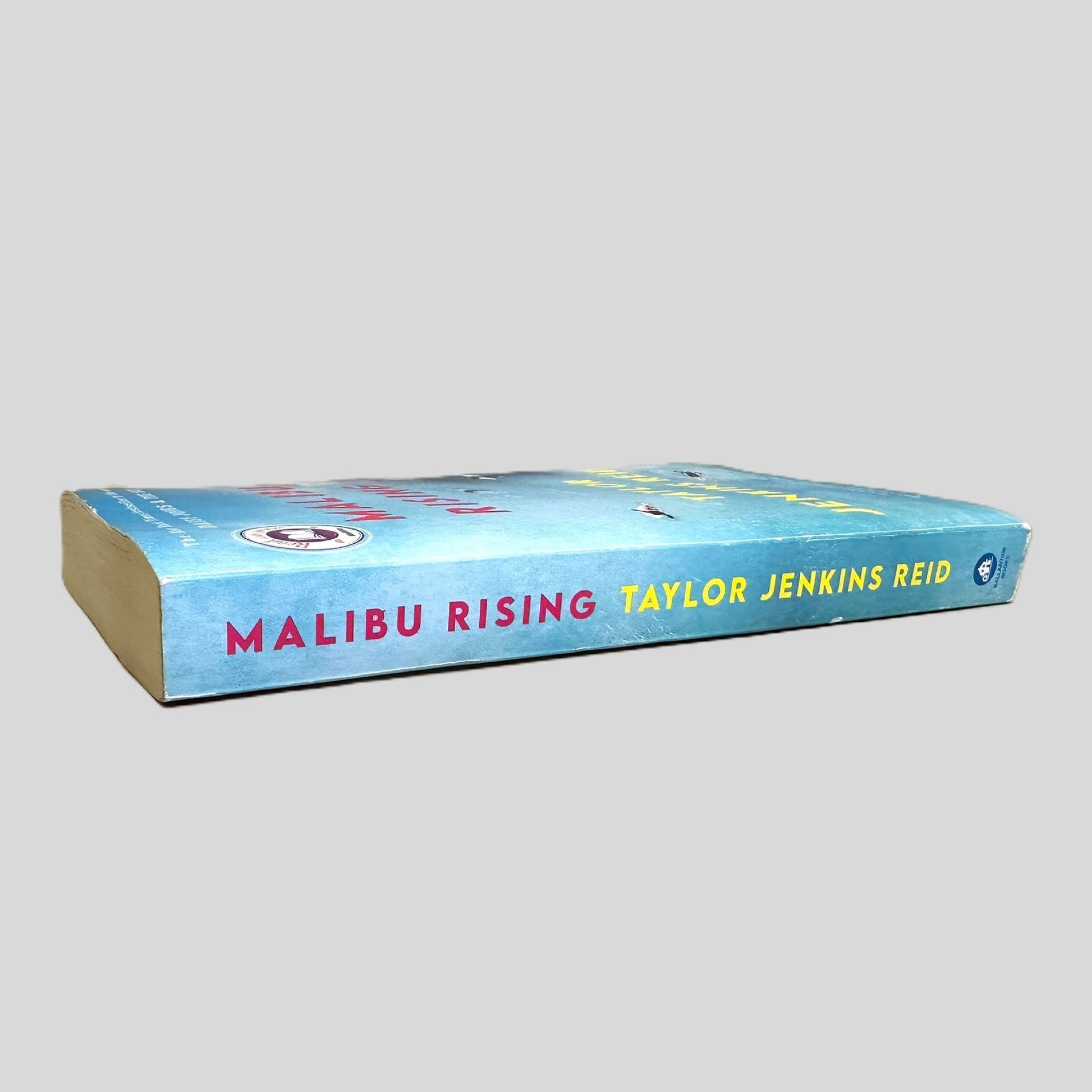 Side-angle view of 'Malibu Rising' by Taylor Jenkins Reid resting on a neutral background. The screen captures the striking red and white typography and Ballantine Books logo on the spine, reflecting the high-quality design of this New York Times bestseller. The layout emphasizes the surf-culture aesthetic of the novel, showcasing the sharp corners.