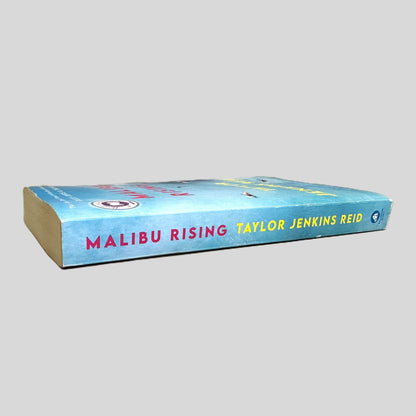 Side-angle view of 'Malibu Rising' by Taylor Jenkins Reid resting on a neutral background. The screen captures the striking red and white typography and Ballantine Books logo on the spine, reflecting the high-quality design of this New York Times bestseller. The layout emphasizes the surf-culture aesthetic of the novel, showcasing the sharp corners.