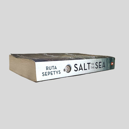 Side-angle view of 'Salt to the Sea' by Ruta Sepetys resting on a neutral background. The screen captures the striking black typography and Penguin logo on the spine, reflecting the high-quality design of this bestselling trade paperback. The layout emphasizes the literary significance of the work, showcasing the sharp corners and authentic condition of this modern historical classic.