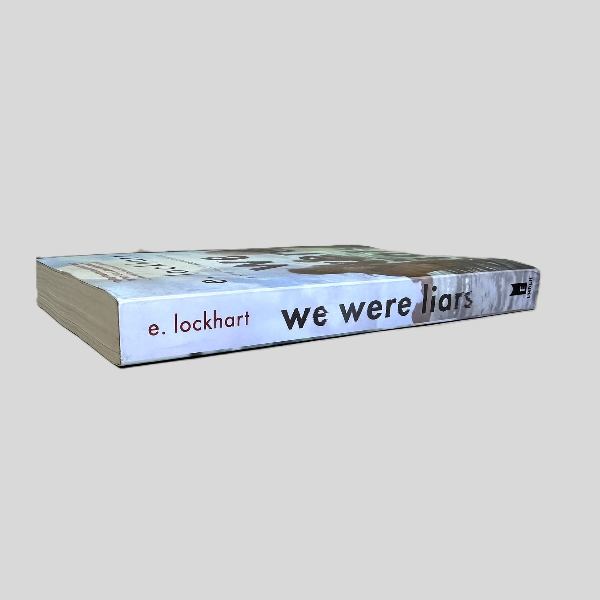 Side-angle view of 'We Were Liars' by E. Lockhart resting on a dark surface. The screen captures the iconic typography and Ember logo on the spine, reflecting the notable design by Associate Art Director Angela Carlino. The layout emphasizes the atmospheric New England setting, showcasing the sharp corners and authentic cover wrap of this critically acclaimed YA bestseller.