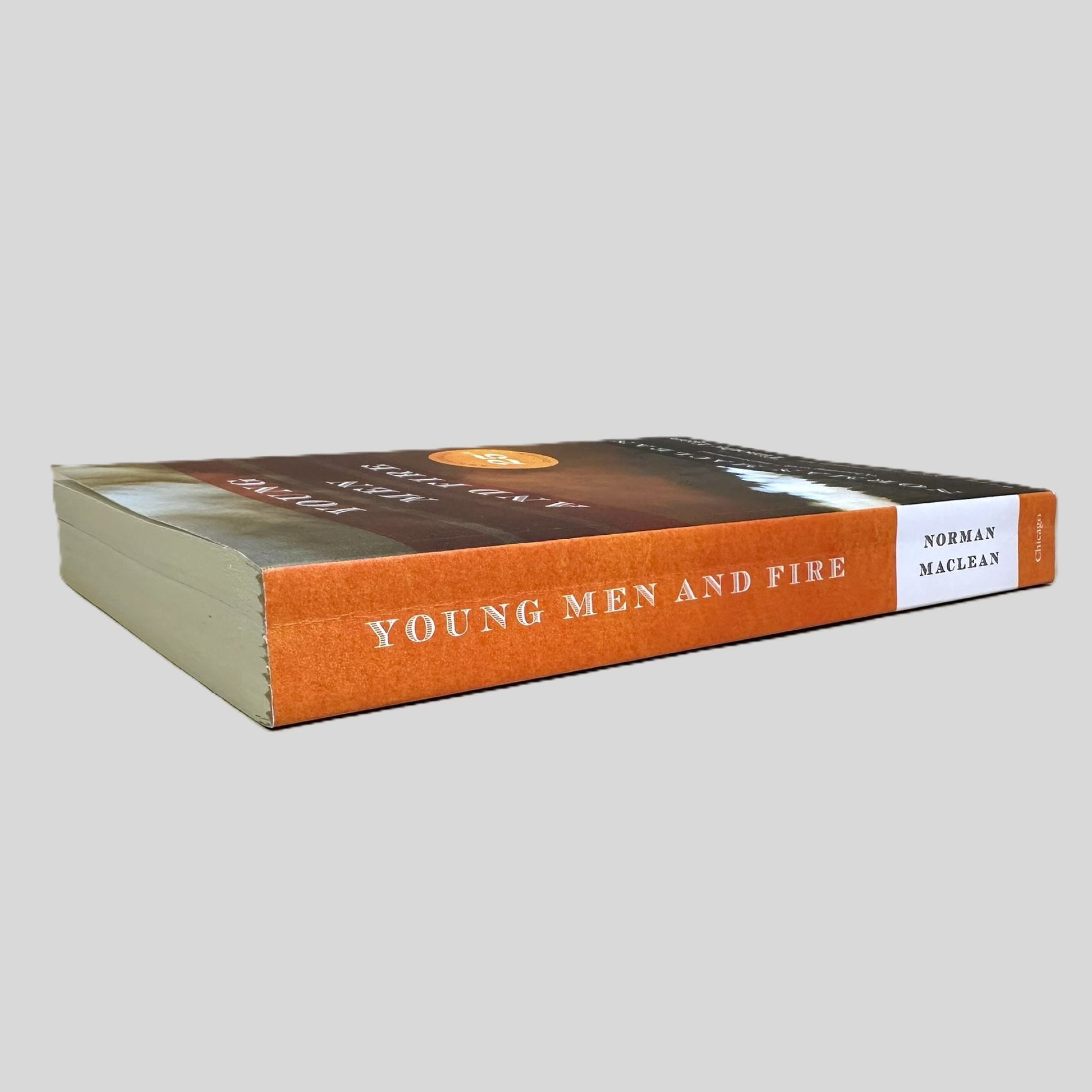 Side-angle view of 'Young Men and Fire' by Norman Maclean resting on a neutral background. The screen captures the bold title and author name on the orange spine, reflecting the distinguished academic press design of the University of Chicago. The layout emphasizes the literary importance of this renowned non-fiction account, showcasing the substantial page count and pristine edges.