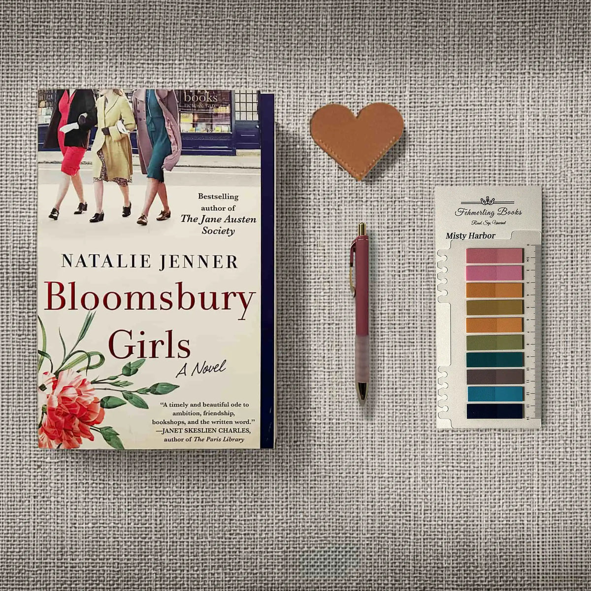 A styled flat-lay featuring the book 'Bloomsbury Girls' by Natalie Jenner, a pink pen, a small heart-shaped leather bookmark, and Misty Harbor annotation tabs from Fehmerling Books, all arranged on a textured gray fabric background.
