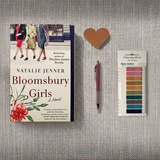 A styled flat-lay featuring the book 'Bloomsbury Girls' by Natalie Jenner, a pink pen, a small heart-shaped leather bookmark, and Misty Harbor annotation tabs from Fehmerling Books, all arranged on a textured gray fabric background.