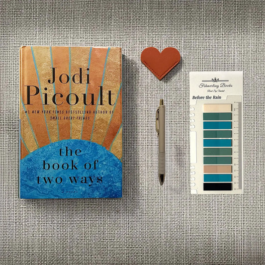 A styled flat-lay featuring the book 'The Book of Two Ways' by Jodi Picoult, a pen, a leather heart-shaped bookmark, and Before the Rain annotation tabs from Fehmerling Books on a textured gray fabric background.