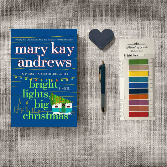 A styled flat-lay featuring the book 'Bright Lights, Big Christmas' by Mary Kay Andrews, a dark blue heart-shaped bookmark, a black pen, and Bright Idea annotation tabs, all arranged on a textured gray linen background.