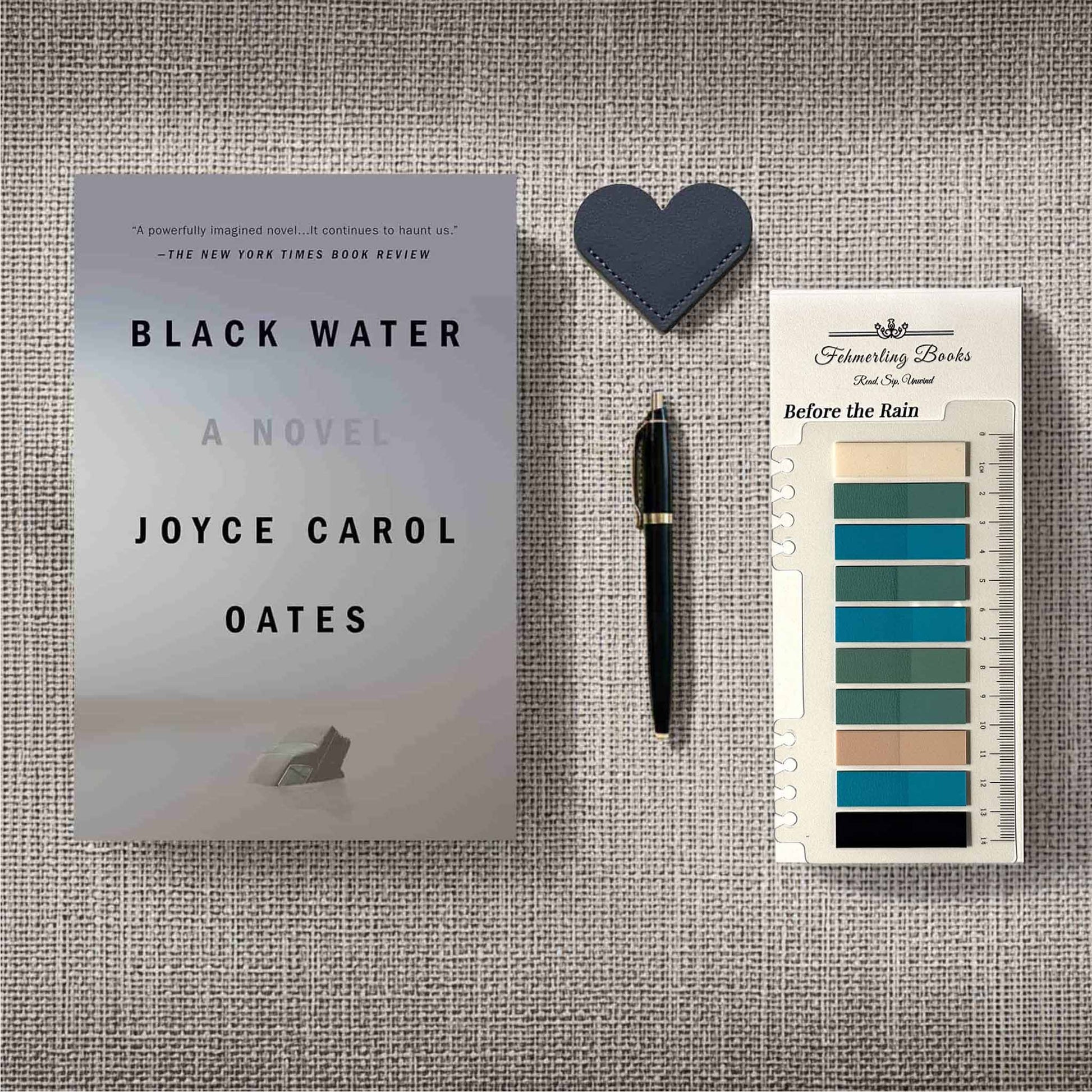A styled flat-lay featuring the book 'Black Water' by Joyce Carol Oates, a black pen, a dark blue heart-shaped leather bookmark and Before the Rain annotation tabs on a textured gray linen background.