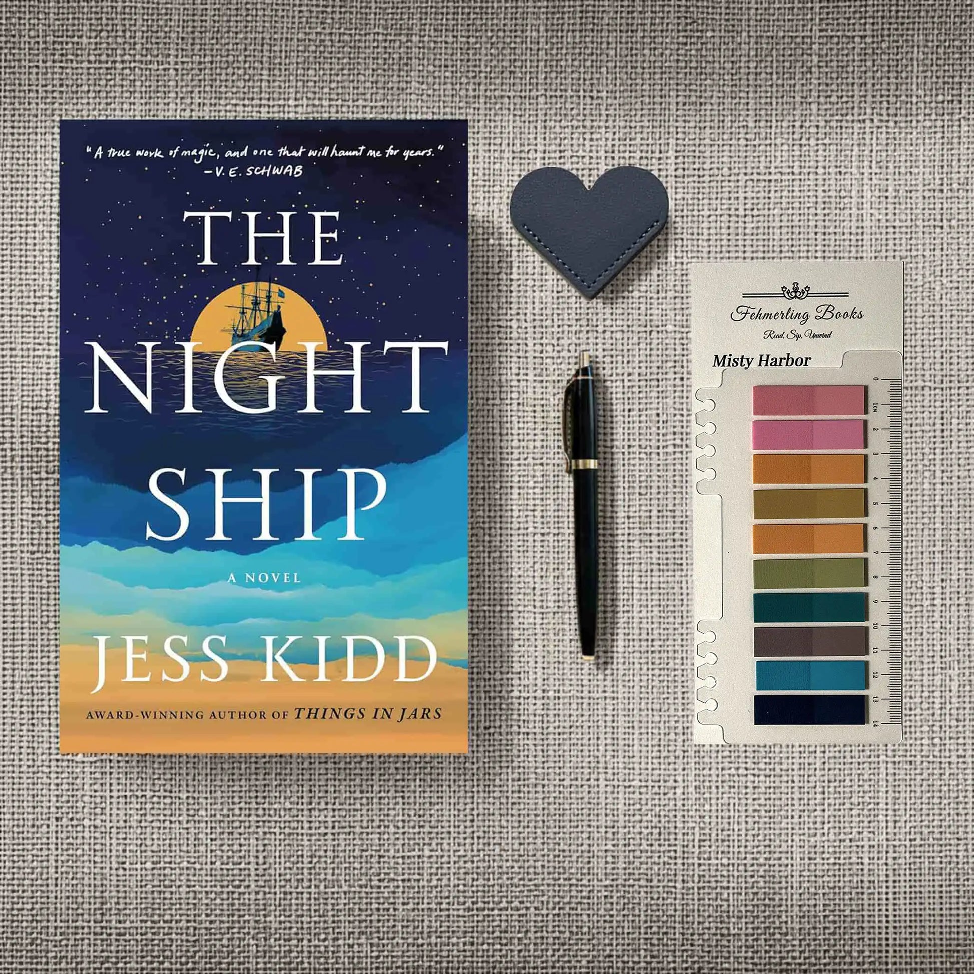 A styled flat-lay featuring the book 'The Night Ship' by Jess Kidd, a black pen, a dark blue heart-shaped leather bookmark, and a card of annotation tabs from 'Fehmerling Books' titled 'Misty Harbor', all arranged on a textured gray linen background.