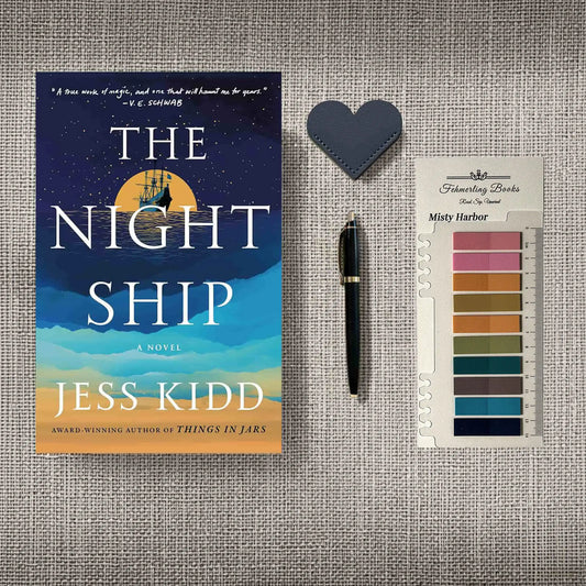 A styled flat-lay featuring the book 'The Night Ship' by Jess Kidd, a black pen, a dark blue heart-shaped leather bookmark, and a card of annotation tabs from 'Fehmerling Books' titled 'Misty Harbor', all arranged on a textured gray linen background.