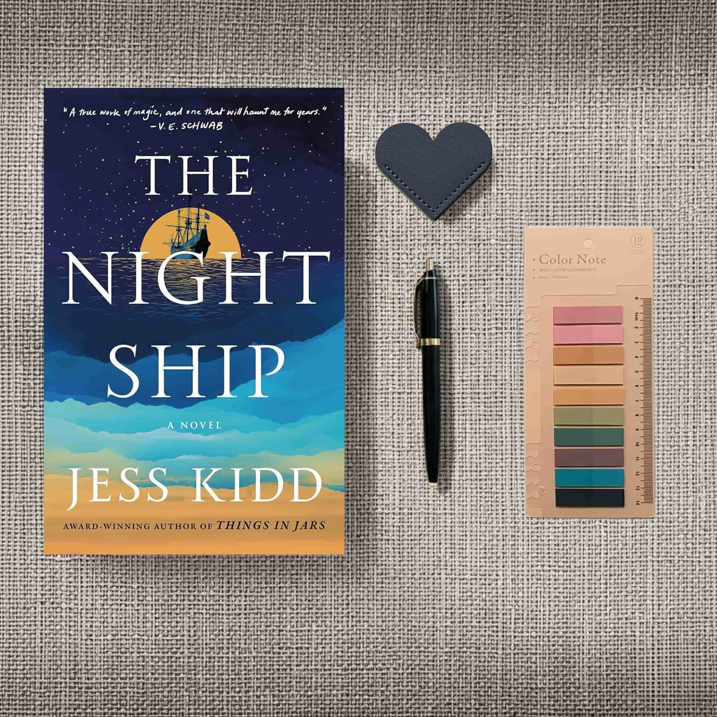 A flat-lay product image for the "Bookworm Bundle" featuring a New condition paperback copy of "The Night Ship" by Jess Kidd. The bundle is arranged on a gray textured background and includes a black and gold pen, a dark blue heart-shaped leather bookmark, and a pack of translucent annotation tabs in muted earth tones.