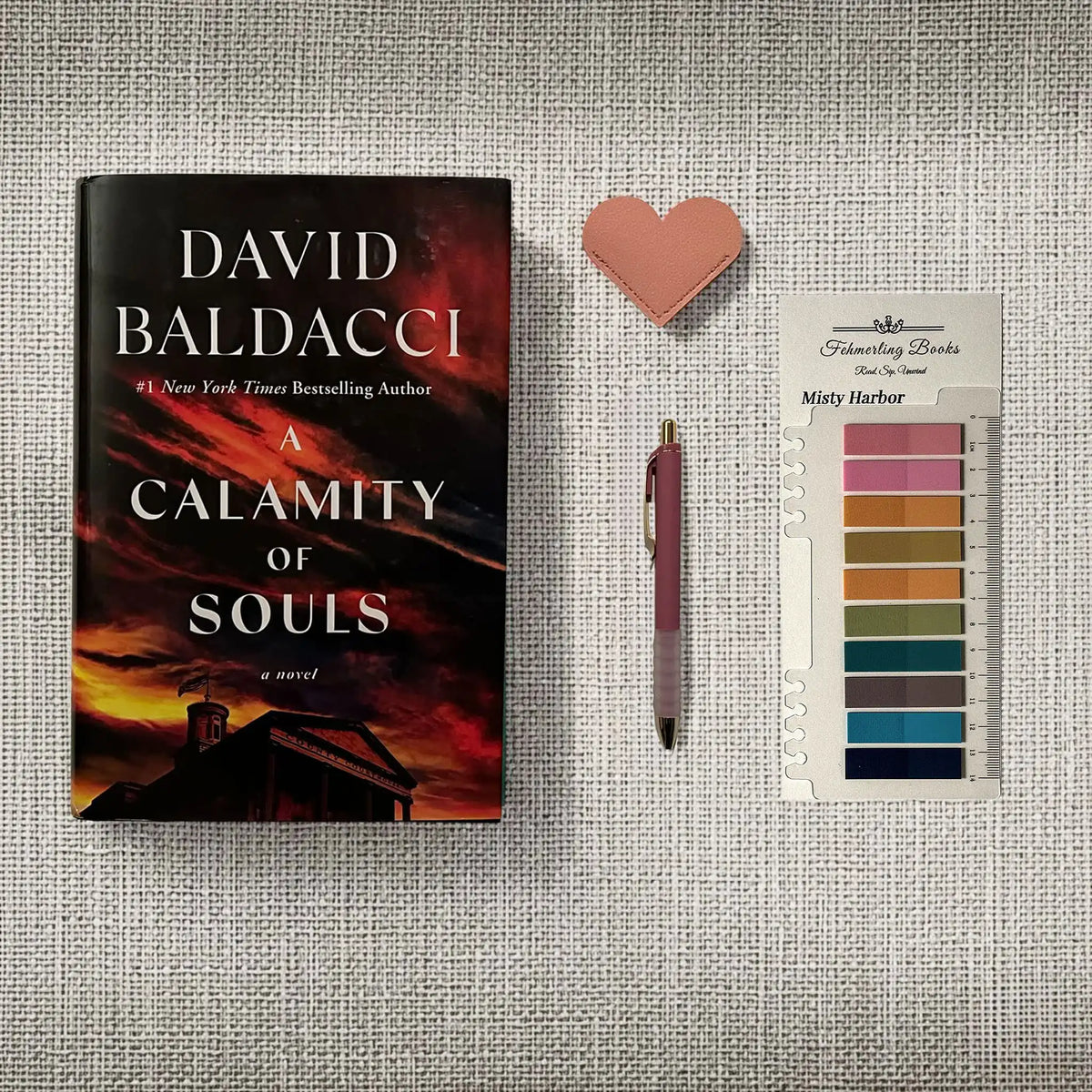 A book titled 'A Calamity of Souls' by David Baldacci is displayed on a textured gray fabric background alongside a pink heart-shaped leather bookmark, a purple pen, and Misty Harbor annotation tabs.