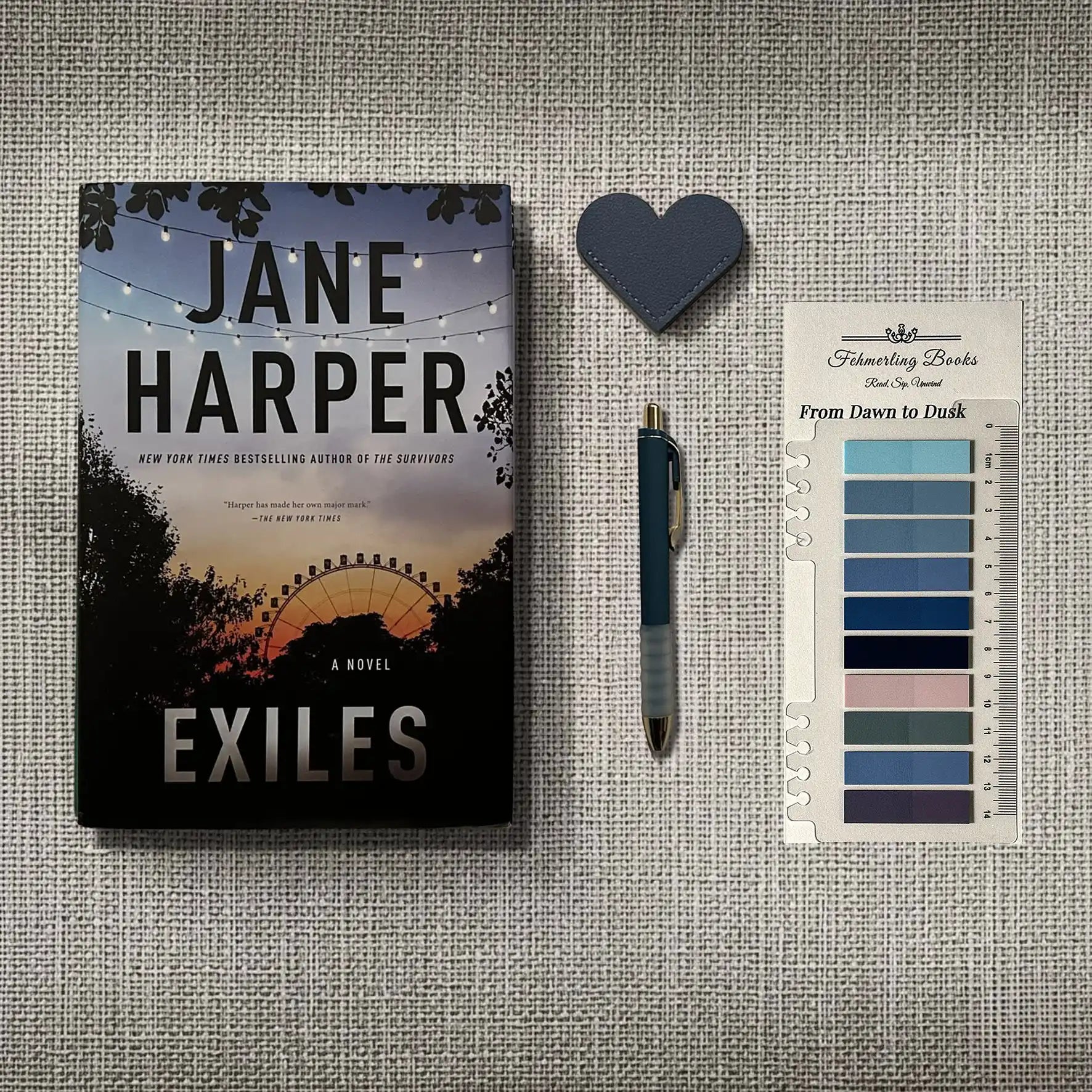 A book titled "Exiles" by Jane Harper is displayed on a textured gray fabric background alongside a dark blue heart-shaped object, a blue pen, and From Dawn to Dusk annotation tabs from Fehmerling Books.