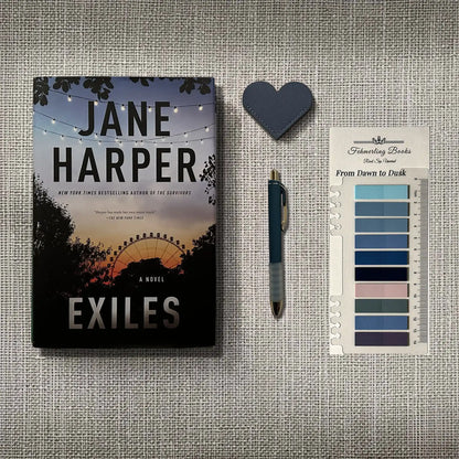 A book titled "Exiles" by Jane Harper is displayed on a textured gray fabric background alongside a dark blue heart-shaped object, a blue pen, and From Dawn to Dusk annotation tabs from Fehmerling Books.