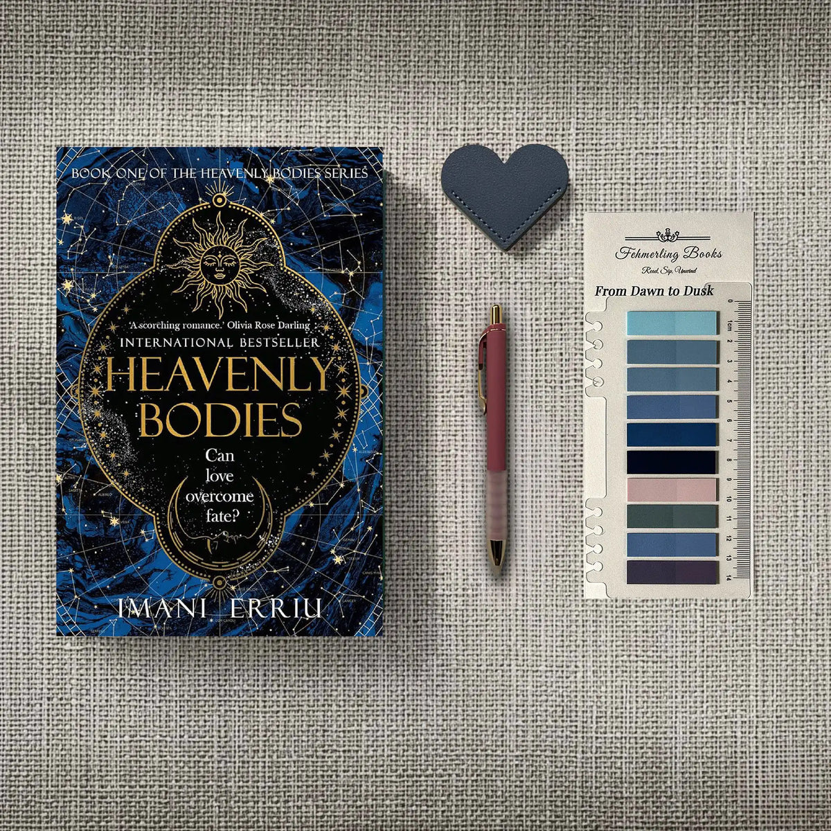 A styled flat-lay featuring the book 'Heavenly Bodies' by Imani Erriu, a dark blue heart-shaped leather bookmark, a pink pen, and 'From Dawn to Dusk' annotation tabs, all arranged on a textured gray linen background.