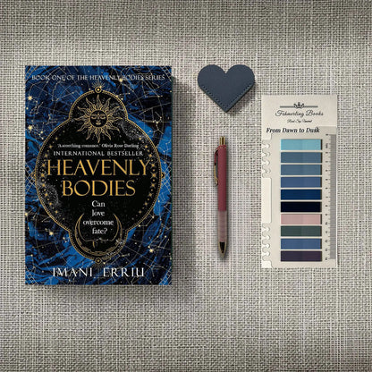 A styled flat-lay featuring the book 'Heavenly Bodies' by Imani Erriu, a dark blue heart-shaped leather bookmark, a pink pen, and 'From Dawn to Dusk' annotation tabs, all arranged on a textured gray linen background.