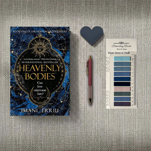 A styled flat-lay featuring the book 'Heavenly Bodies' by Imani Erriu, a dark blue heart-shaped leather bookmark, a pink pen, and 'From Dawn to Dusk' annotation tabs, all arranged on a textured gray linen background.