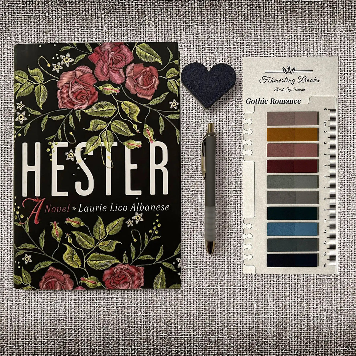 A book titled 'HESTER' by Laurie Lico Albanese is displayed on a textured gray fabric background alongside a dark blue heart-shaped bookmark, a gray pen, and Gothic Romance annotation tabs from Fehmerling Books.