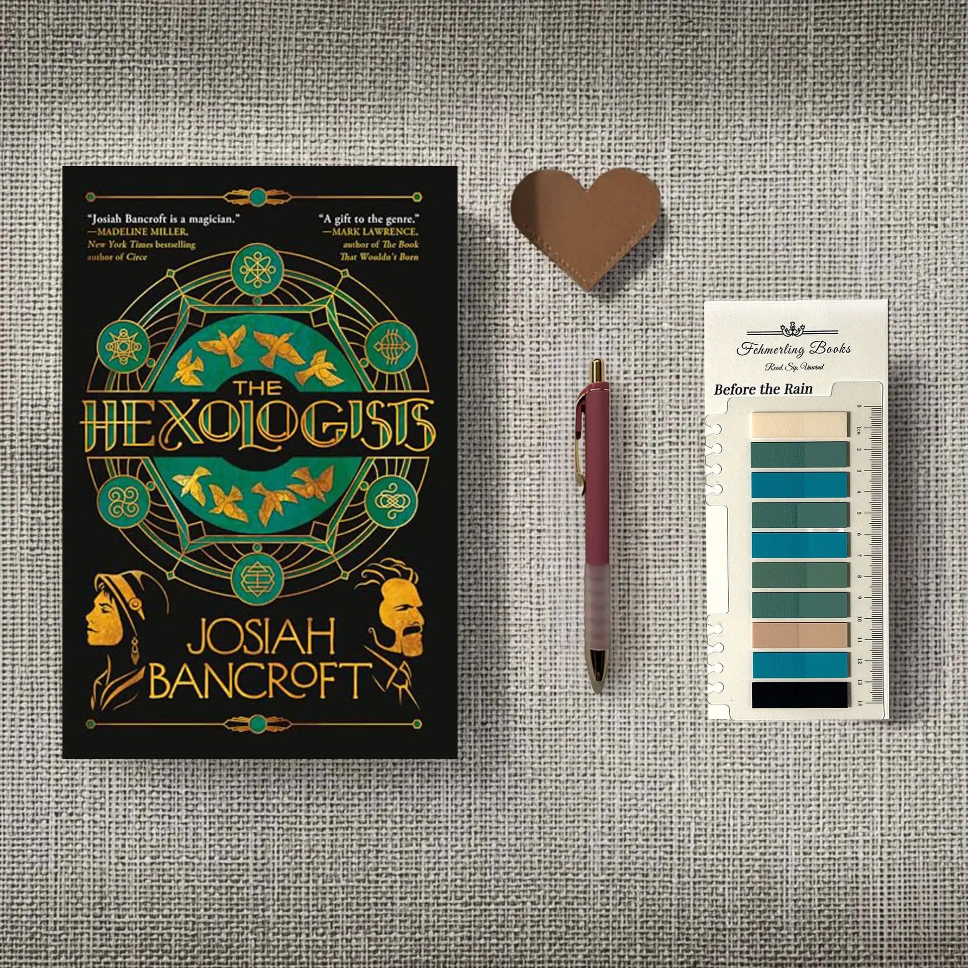 A styled flat-lay featuring the book cover of 'The Hexologis' by Josiah Bancroft, a heart-shaped leather bookmark, a maroon pen, and 'Before the Rain' annotation tabs from Fehmerling Books, all arranged on a textured gray fabric background.