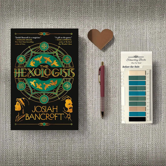 A styled flat-lay featuring the book cover of 'The Hexologis' by Josiah Bancroft, a heart-shaped leather bookmark, a maroon pen, and 'Before the Rain' annotation tabs from Fehmerling Books, all arranged on a textured gray fabric background.