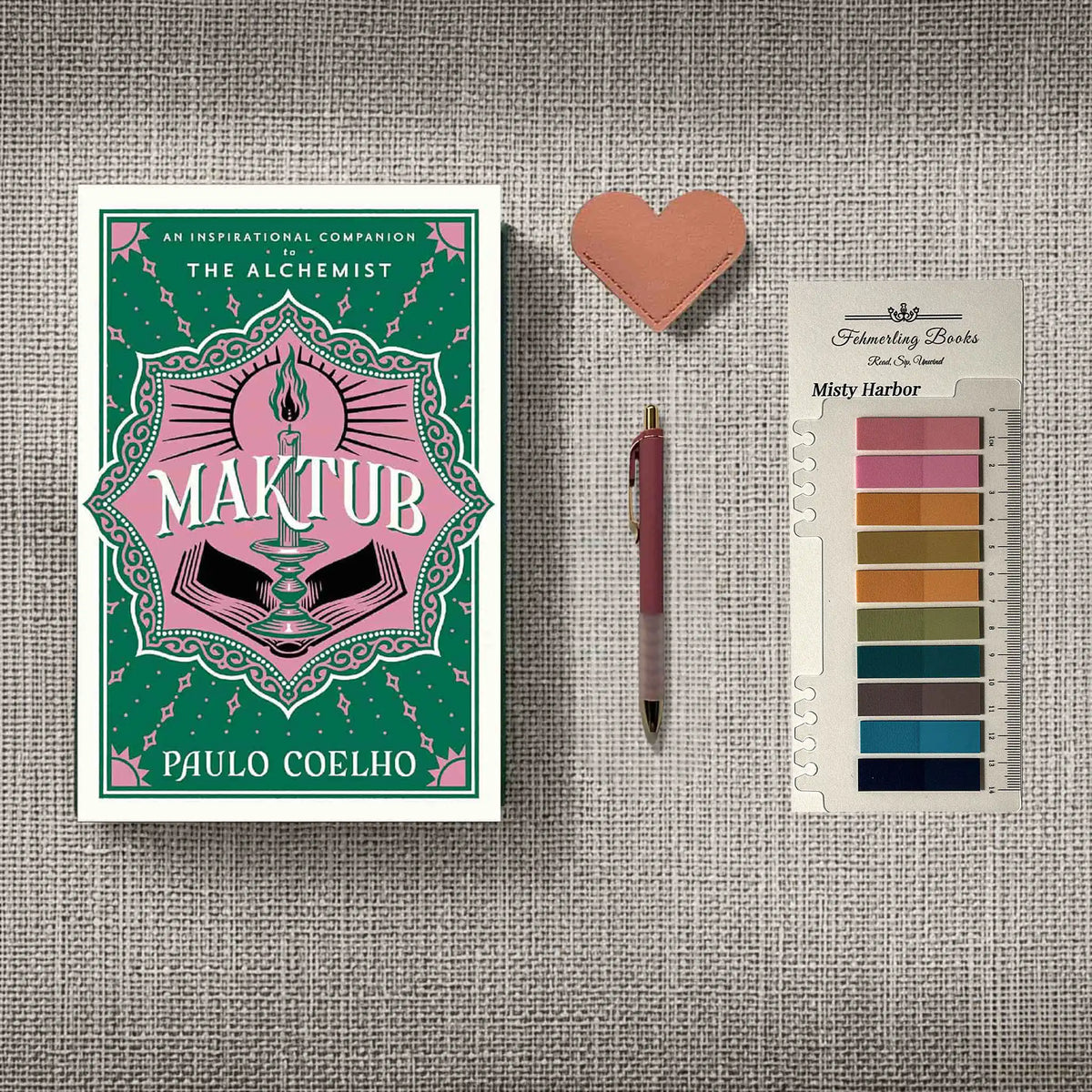 A styled flat-lay featuring the book 'Maktub' by Paulo Coelho, a pink heart-shaped leather bookmark, a pink pen, and Misty Harbor annotation tabs from 'Fehmerling Books' , all arranged on a textured gray fabric background.