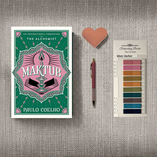 A styled flat-lay featuring the book 'Maktub' by Paulo Coelho, a pink heart-shaped leather bookmark, a pink pen, and Misty Harbor annotation tabs from 'Fehmerling Books' , all arranged on a textured gray fabric background.