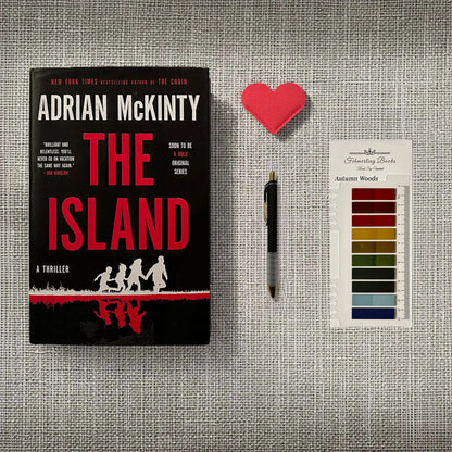 A book titled "The Island" by Adrian McKinty is displayed on a textured gray fabric background, alongside a heart-shaped leather bookmark,  a black pen, and Autumn Woods annotation tabs from Fehmerling Books.