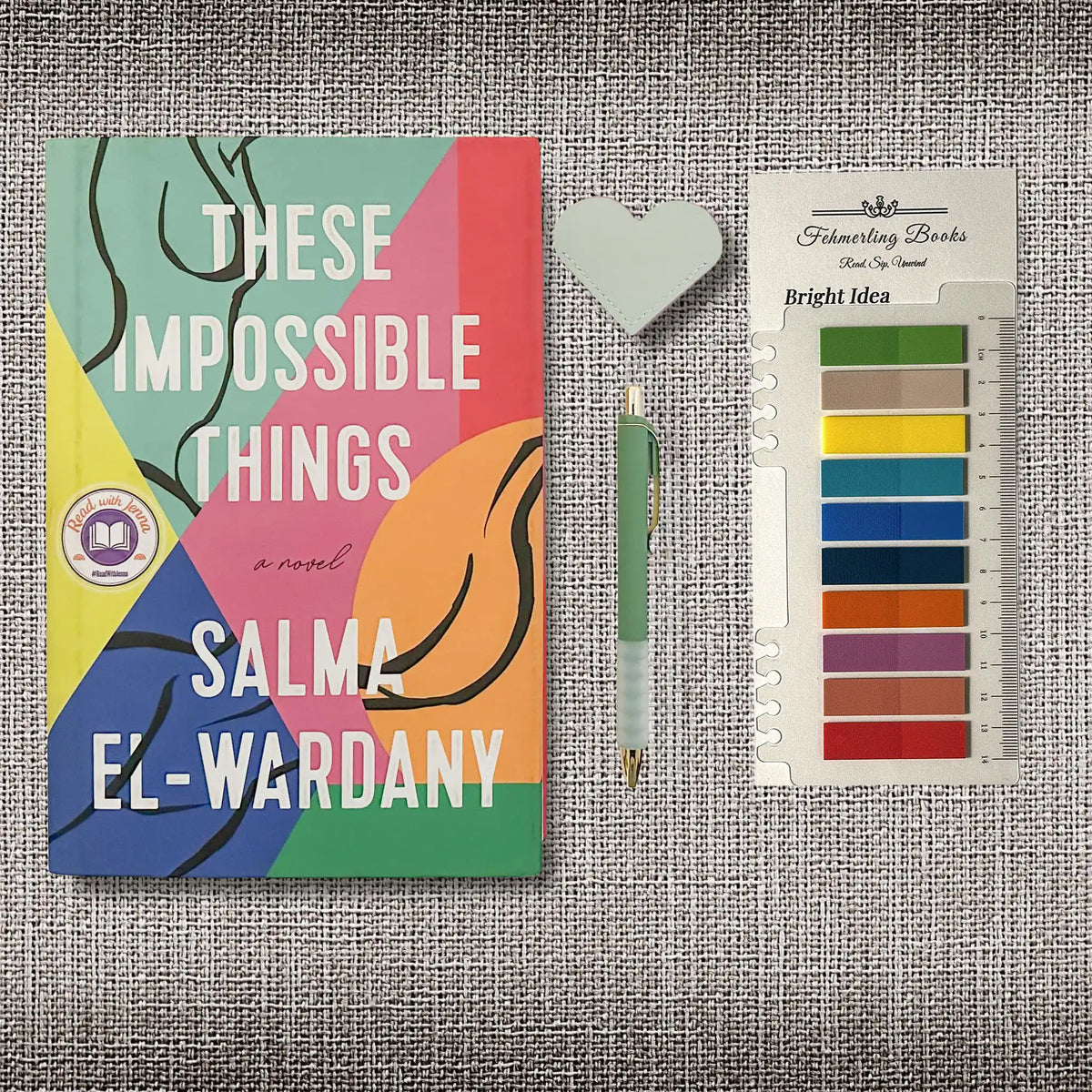 A styled flat-lay featuring the book cover of 'These Impossible Things' by Salma El-Wardany, a pen, a heart shaped leather bookmark, and Bright Idea annotation tabs from Fehmerling Books, all arranged on a textured gray fabric background.